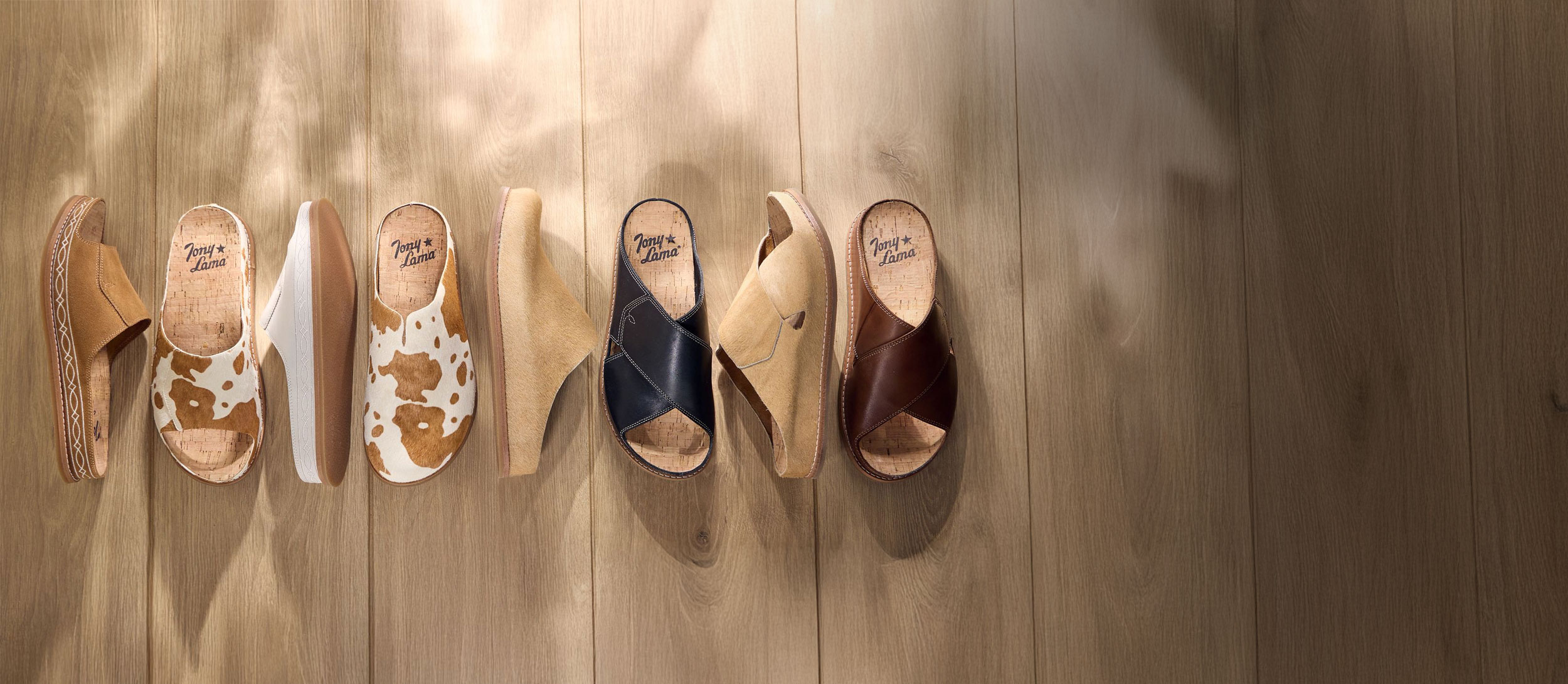 A lineup of the Playa sandal and clog collection in various colors on a hardwood floor.